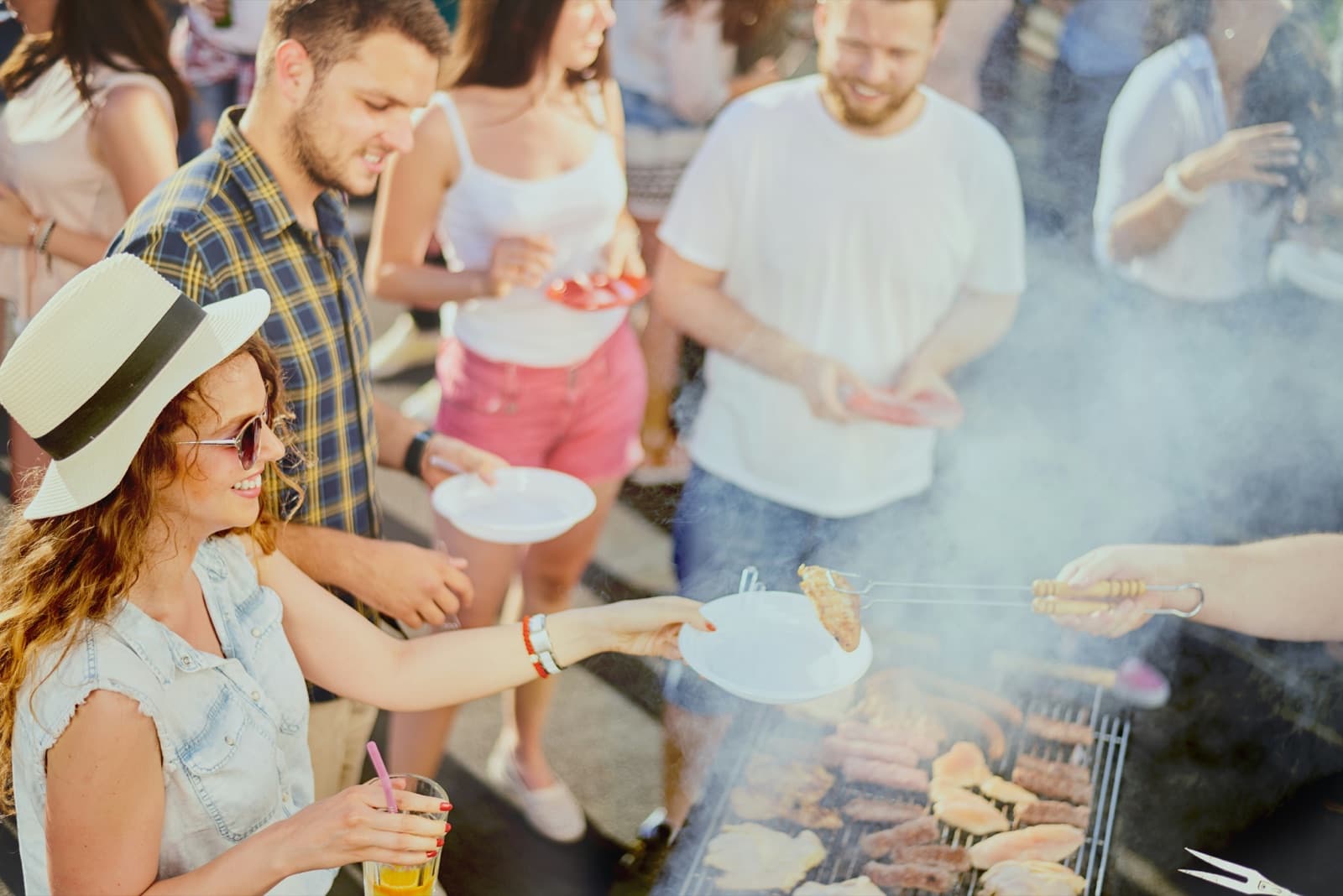 Vänner i sommarkläder samlas kring en rykande grill och delar mat utomhus