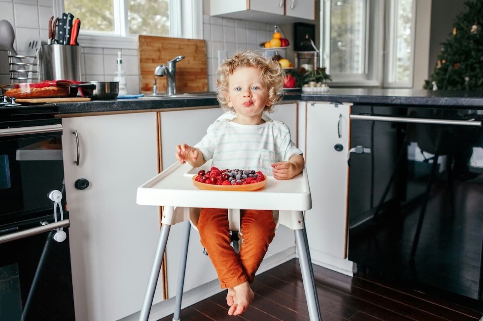 Ett litet barn sitter i barnstol och äter bär i ett kök
