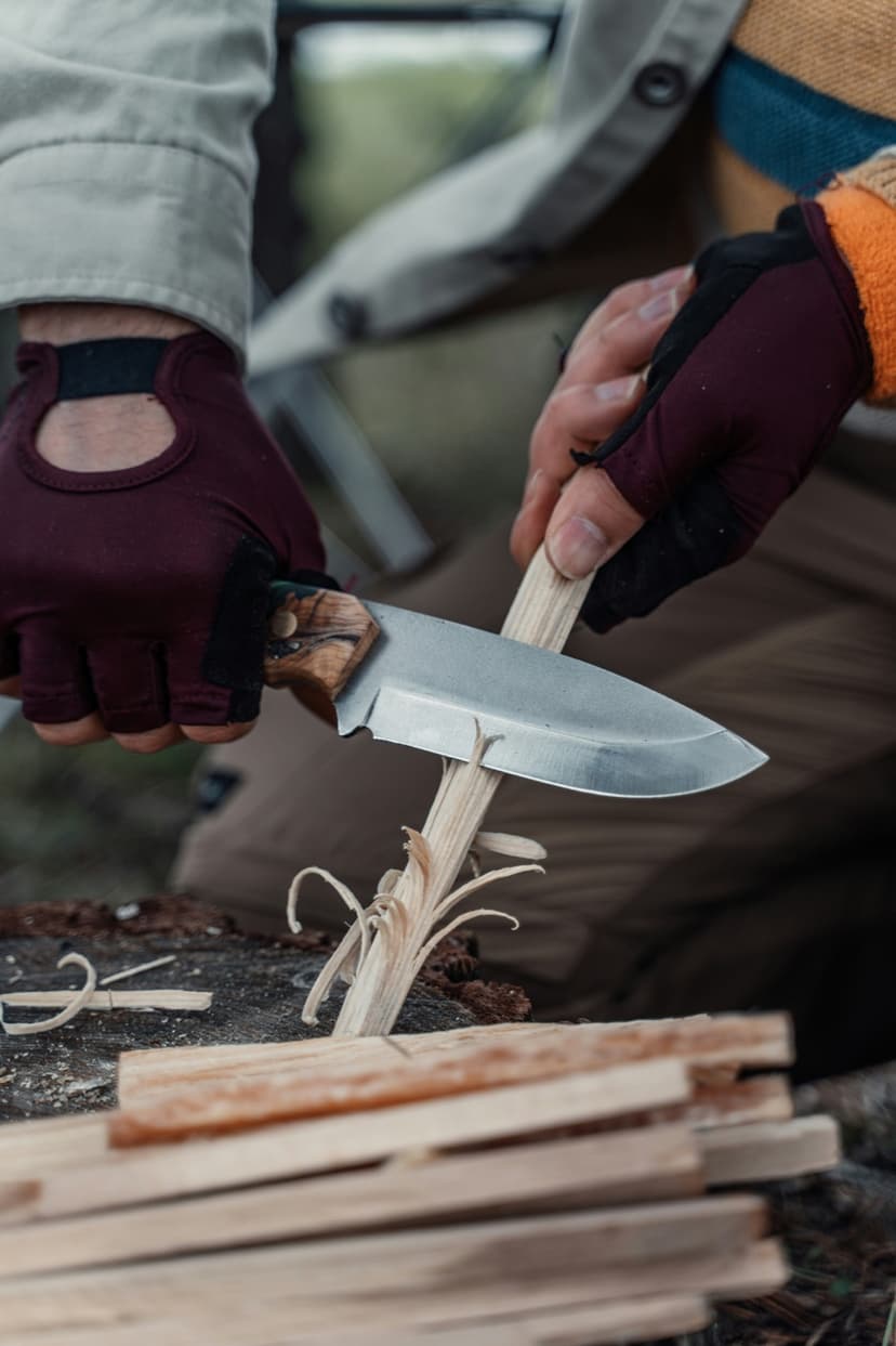 Närbild på händer i fingerlösa handskar som täljer en träpinne med kniv