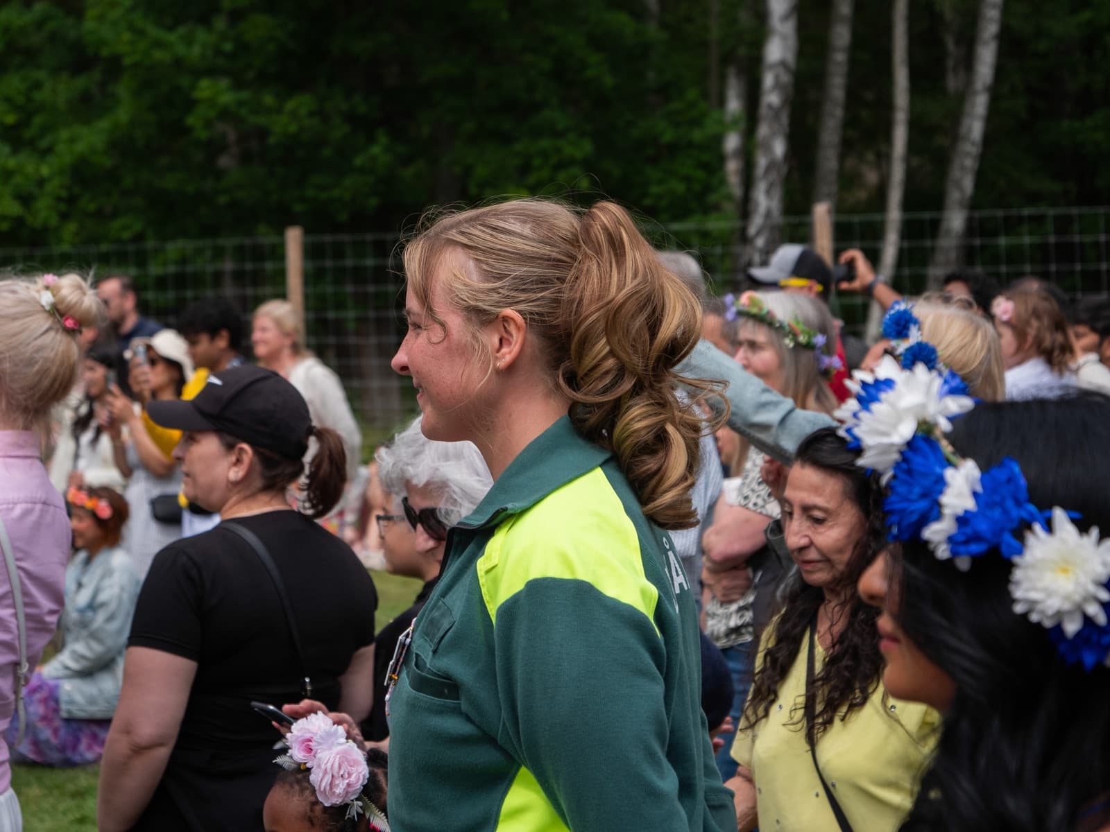 En person i grön och gul larmklädsel står bland festklädda besökare med blomsterkransar i håret