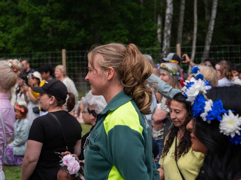 En person i grön och gul larmklädsel står bland festklädda besökare med blomsterkransar i håret