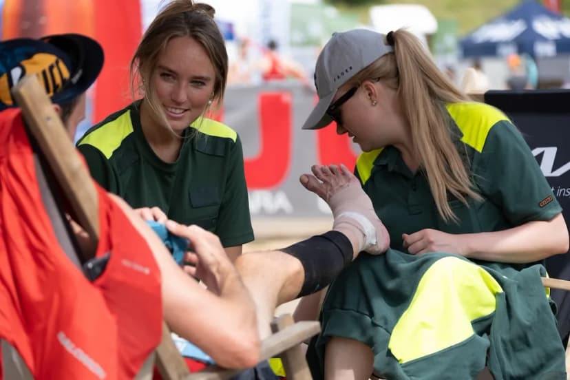 Två sjukvårdare i grön-gul uniform lindar om en handled på en deltagare vid ett utomhusevenemang