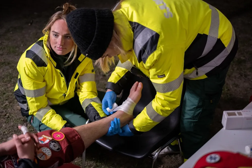 Två personer i gulgröna reflexjackor lindar om en fot på en sittande person utomhus i mörker