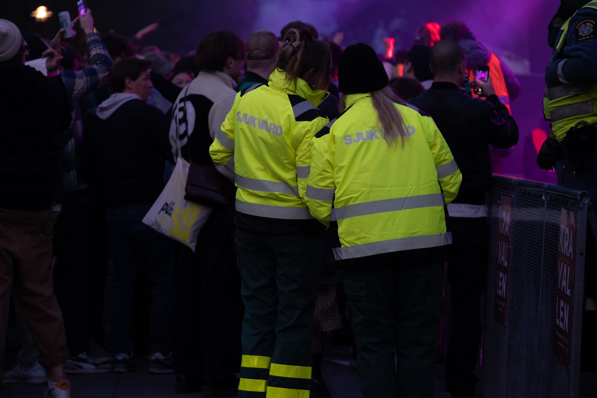 Två sjukvårdare i varseljacka står vid avspärrningen framför scenen under ett evenemang