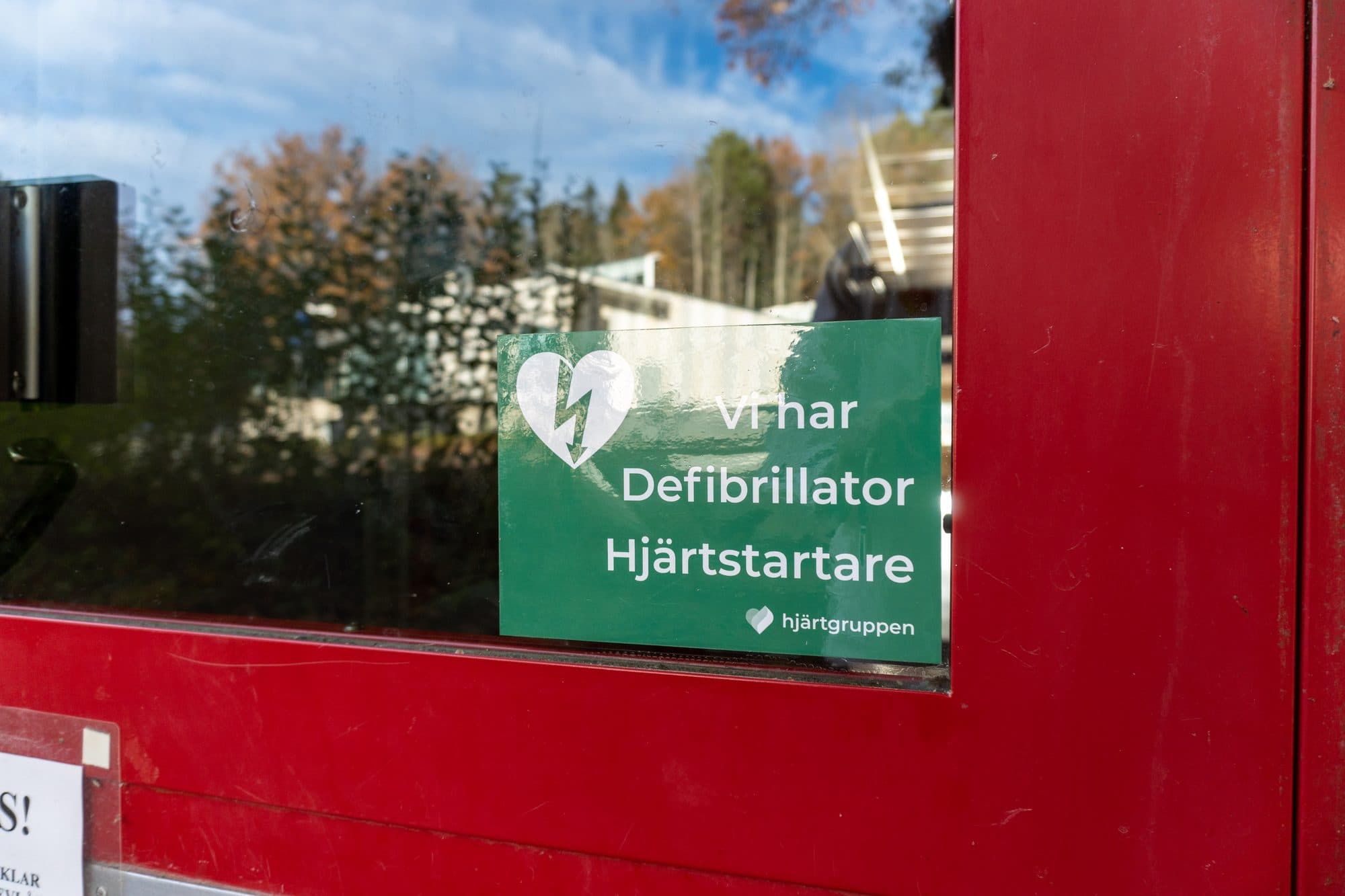Grön skylt med texten Vi har defibrillator hjärtstartare på en röd entrédörr
