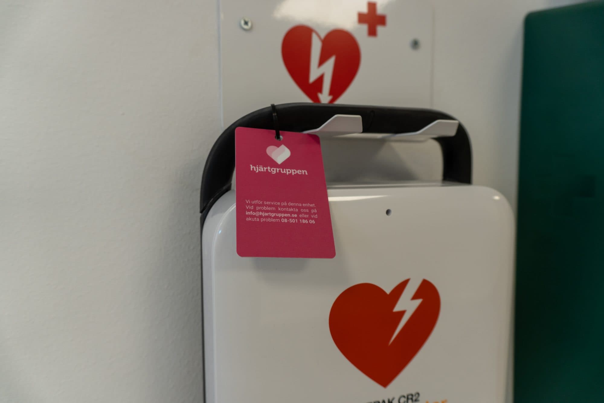 Vit hjärtstartare på vägg med Hjärtgruppens servicebricka på handtaget